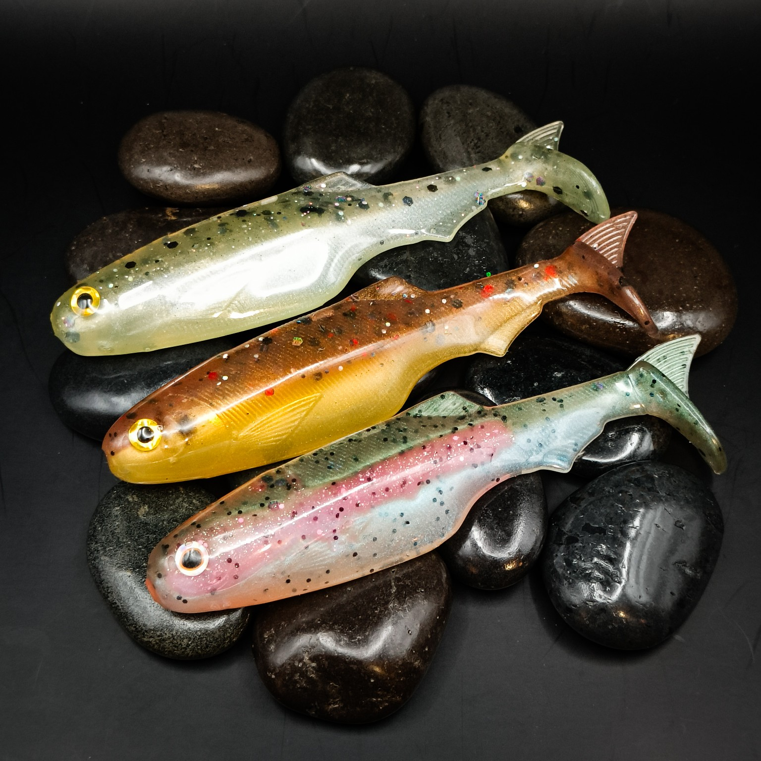 Three Premium Trout Series Scrapper lures in Stockie, Colne Gold and Steel Rose, arranged on dark pebbles.