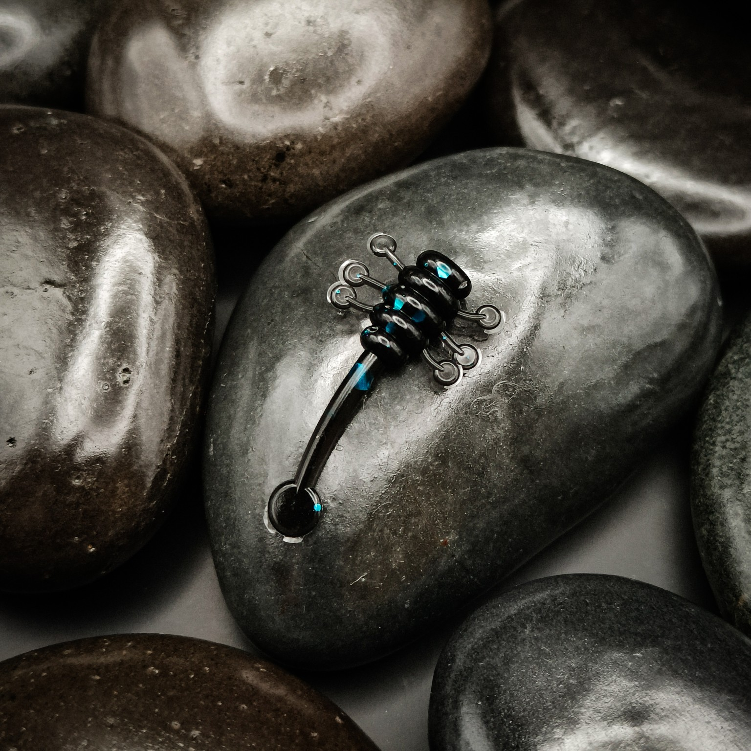 Sting Tail 2.8 cm in Black & Blue, with a black ribbed body, blue glitter, fine side appendages and a long tail, displayed on dark pebbles.